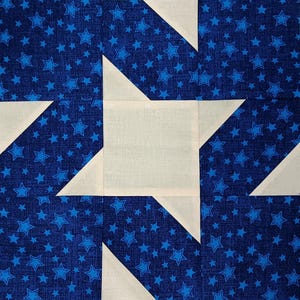 May include: Close-up of a quilt block featuring a star pattern. The design includes a central white square with white triangles and a blue background filled with blue stars. The fabric is likely cotton.