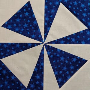 May include: A quilt block with a pinwheel design. The block is made of white fabric and blue fabric with white stars. The blue fabric is arranged in a pinwheel pattern with the white fabric in between each section.