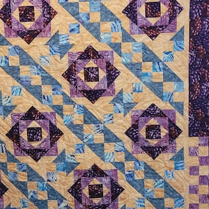 May include: A patterned quilt with a repeating design of squares and diamonds in beige, blue, and purple. The textile features geometric shapes and a textured finish, ideal for home furnishings.