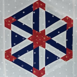May include: A geometric quilt block featuring a starburst design. The design incorporates red, white, and blue fabrics. The blue sections have white stars. The background is white with small white polka dots. The quilt block is a hexagon shape.
