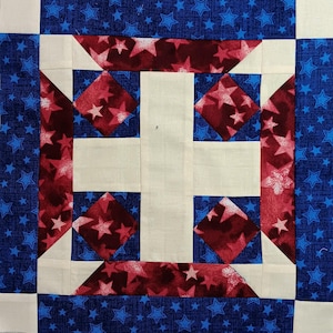 May include: A quilt block with a white cross in the center, surrounded by red and blue fabric with a star pattern. The outer border is blue with white stars.