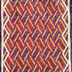 May include: A quilt featuring a geometric design of white, gold and purple stripes. The stripes are arranged diagonally, creating a woven effect.