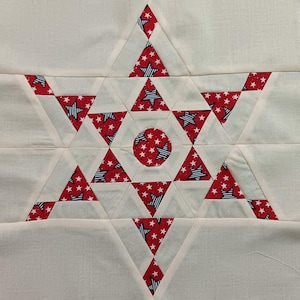 May include: A white fabric background with a red and blue star-shaped quilt block. The quilt block is made up of triangles with a red background and white stars, and triangles with a blue and white striped background and white stars. The center of the quilt block has a circle with a red background and white stars.