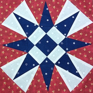 Op de afbeelding: Een vierkant stoffen blok met een sterrenpatroon. De centrale ster is samengesteld uit blauwe stof met witte sterren en wit-blauwe geruite vierkanten. De achtergrond is rood met gouden sterren, wat een patriottisch thema creëert.