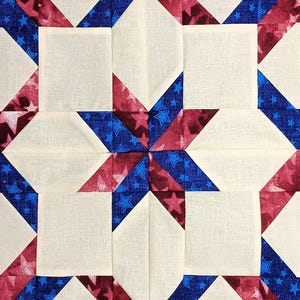 May include: Close-up of a quilt block with a starburst design. The block features a cream-colored square in the center, surrounded by red and blue star-patterned fabric. The design is reminiscent of the American flag.
