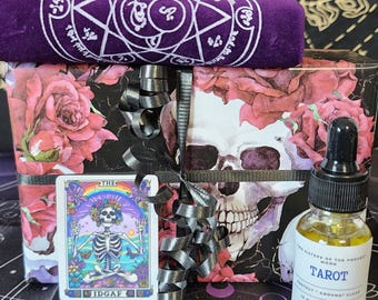 Könnte beinhalten: Ein Geschenkset mit Tarotkarte, Ölflasche und einem gerollten lila Tuch mit weißem Pentagramm-Design. Die Geschenkbox ist in Papier mit Rosen- und Totenkopfmuster verpackt, mit silbernem Band gebunden und auf einem schwarzen Tuch platziert.
