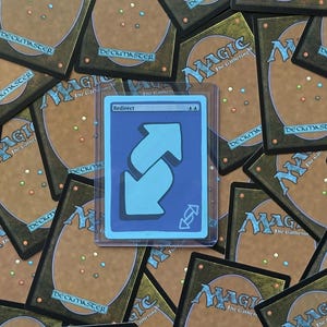Puede incluir: Una carta azul de Magic: The Gathering con la palabra "Redirect" y dos flechas celeste claro que apuntan en direcciones opuestas. La carta está en una funda protectora, rodeada de otras cartas de Magic: The Gathering.