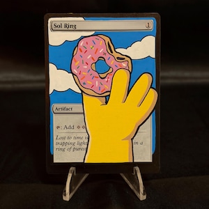 May include: A custom trading card titled Sol Ring featuring a hand-painted illustration of a yellow four-fingered hand holding a pink frosted donut with colorful sprinkles against a blue sky with white clouds. The card text includes Artifact and Add.
