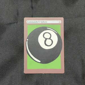 May include: A card with the text "Commander's Sphere" and a large black eight ball on a green background. The card has a brown border and is set against a dark gray background.
