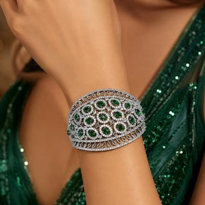 Könnte beinhalten: Ein breites Armband mit einer Basis aus silberfarbenem Metall und einem komplexen, durchbrochenen Gitterdesign. Das Schmuckstück ist mit zahlreichen oval geschliffenen smaragdgrünen Steinen und kleinen klaren weißen Kristallen besetzt.