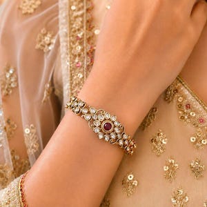 Peut inclure: Un bracelet doré orné d'une pierre rouge ronde centrale entourée d'un groupe de pierres claires en forme de goutte, serties dans une monture métallique complexe. Le bracelet est porté au poignet sur un vêtement beige brodé d'or.