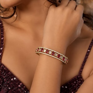 Puede incluir: Un brazalete rígido articulado en tono dorado que presenta una fila central de piedras rojas de corte ovalado alternadas con piedras claras de corte baguette, bordeado por dos filas delgadas de pequeñas piedras claras en pavé.
