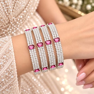May include: Four silver-toned bangles featuring three rows of small clear pavé stones and a single rectangular fuchsia-coloured centre stone. The bracelets are worn together on the wrist, showcasing a metallic finish and vibrant pink accents.
