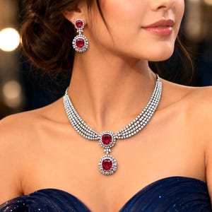 May include: A silver-toned necklace and matching drop earring set featuring cushion-cut red gemstones surrounded by halos of small clear stones. The necklace consists of four rows of clear stones leading to a central red stone and a dangling pendant.