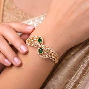 May include: A gold-toned open cuff bracelet featuring two teardrop-shaped ends, each centred with an oval-cut green stone. The metal surface is intricately detailed with a filigree pattern and embedded with small, clear, sparkling stones.