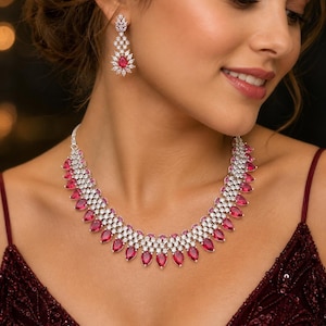 Puede incluir: Un deslumbrante conjunto de joyas con collar y pendientes a juego. El collar y los pendientes muestran una combinación de piedras preciosas claras y de color rojo rubí. El collar es de estilo gargantilla, y los pendientes son colgantes.