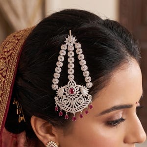 May include: A woman wearing a diamond and ruby bridal jewelry set. The set includes a headpiece, earrings, and a necklace. The headpiece is a detailed design with dangling elements. The jewelry complements the red and gold embroidered fabric.
