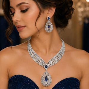 Può includere: Un set composto da collana e orecchini pendenti color argento con intricati lavori in filigrana e pietre pavé trasparenti. Ogni pezzo è centrato da una pietra preziosa blu a forma di goccia, creando un insieme di gioielli coeso e dettagliato.
