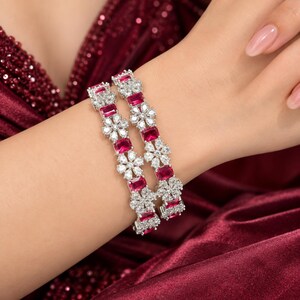 May include: Two silver-toned bangles featuring alternating rectangular red stones and clear floral-shaped stones. The bangles are designed with a repeating pattern of these red and clear elements, creating a continuous decorative band around the wrist.