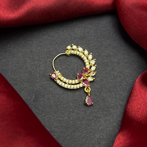 Puede incluir: Un aro de nariz dorado con un diseño de media luna. El anillo está adornado con pequeñas piedras preciosas blancas y rojas. Una sola gema roja en forma de lágrima cuelga de la parte inferior. La joya está sobre una superficie negra y tela roja.