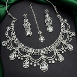 Op de afbeelding: Een zilverkleurige sieradenset met een ketting, oorbellen en een hoofdband. De set is versierd met heldere, sprankelende edelstenen in verschillende vormen, waaronder druppels en vierkanten. De sieraden worden gepresenteerd tegen een donkere achtergrond.