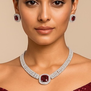 May include: A matching jewelry set featuring a necklace and earrings. The necklace and earrings have a square, deep red center stone surrounded by a halo of small, clear stones. The jewelry is set against a maroon sequined dress.