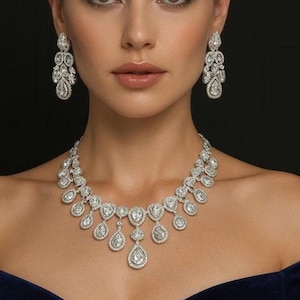 Pode incluir: Uma mulher usando um colar de diamantes e brincos combinando. O colar apresenta pingentes em forma de gota. Os brincos são estilo lustre. A mulher está vestindo um vestido azul escuro com os ombros de fora em um fundo preto.