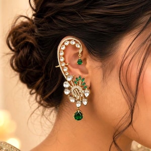 Puede incluir: Un pendiente tipo ear cuff dorado con un diseño curvo adornado con piedras blancas transparentes y pequeños detalles en forma de gota de color verde. La pieza sigue el contorno de la oreja y tiene una piedra verde colgando en la base.