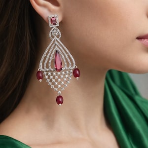 Peut inclure: Boucles d'oreilles pendantes élégantes avec un dessus carré, une grande pierre centrale en forme de goutte et des perles rouges pendantes. Les boucles d'oreilles sont argentées et ornées de petits cristaux étincelants, créant un design luxueux et accrocheur.