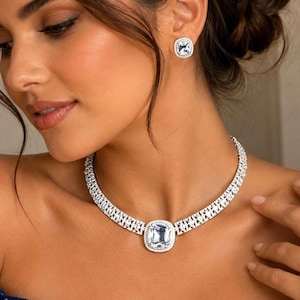 May include: A silver-toned necklace featuring a large, central cushion-cut clear stone surrounded by a halo of smaller stones, connected to a multi-row chain of pavé-set crystals, paired with matching square-shaped stud earrings with clear stones.