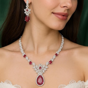 Può includere: Un set di gioielli abbagliante con collana e orecchini abbinati. La collana e gli orecchini sono adornati con scintillanti pietre chiare e vibranti pietre preziose rosse. La collana ha un pendente centrale con una grande pietra rossa a forma di goccia.