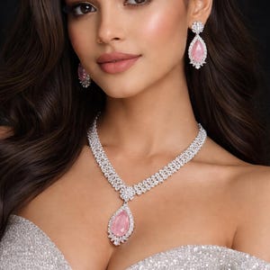 Op de afbeelding: Een schitterende sieradenset met een ketting en oorbellen. De ketting heeft een druppelvormige roze edelsteen hanger, passend bij de oorbellen. De sieraden zijn gezet in een zilverkleurig metaal en hebben ingewikkelde details.