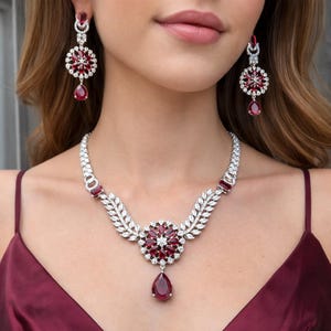 Peut inclure: Un ensemble de bijoux éblouissant comprenant un collier et des boucles d'oreilles assorties. Le collier présente un motif floral central avec un grand pendentif en forme de goutte. Les boucles d'oreilles et le collier sont ornés de pierres rouges et de cristaux clairs, sertis dans un métal argenté.
