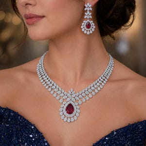 Puede incluir: Un deslumbrante conjunto de collar y pendientes con un gran colgante de rubí en forma de lágrima rodeado de brillantes piedras preciosas blancas. El collar y los pendientes tienen un diseño floral, con un vestido azul oscuro con lentejuelas en el fondo.