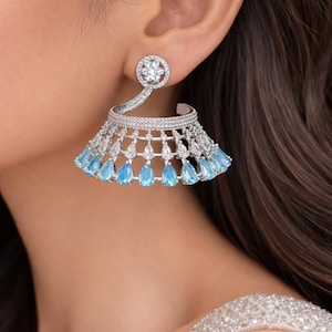 May include: Elegant silver-toned earrings with a circular stud and a cascading design. The earrings feature clear and blue teardrop-shaped gemstones, creating a glamorous and sophisticated look. The earrings are designed to be worn on the ear.