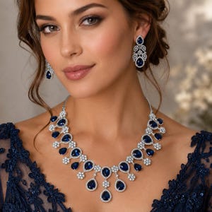 May include: A woman wearing a striking jewellery set. The set includes a necklace and matching earrings. The necklace and earrings feature large, teardrop-shaped, deep blue gemstones surrounded by smaller, clear stones, set in a silver-coloured metal.