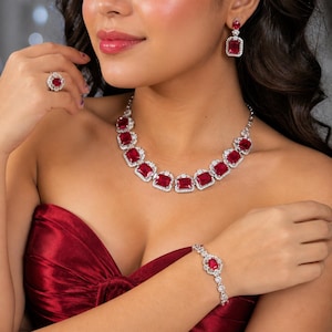Puede incluir: Un conjunto de joyas a juego con un collar, pulsera, anillo y pendientes de color rojo rubí. El collar y la pulsera tienen piedras rojas rectangulares rodeadas de pequeñas piedras transparentes. El anillo y los pendientes tienen una sola piedra roja rectangular.