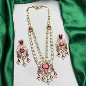 May include: A gold-toned necklace and matching earring set featuring deep red hexagonal and teardrop-shaped stones. The necklace has a long, multi-layered chain design with intricate metalwork and small clear crystals surrounding the red stones.