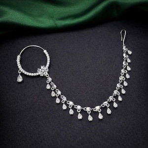 May include: A silver nose ring featuring a floral motif and hanging teardrop crystals. A delicate chain of smaller teardrop crystals extends from the ring. The jewellery is displayed against a dark backdrop, highlighting the intricate design and sparkle.