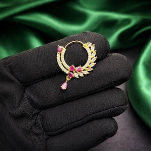 May include: A gold-colored nose ring with a circular design, adorned with clear and pink gemstones. The ring features a teardrop-shaped pink gem pendant and a leaf-like pattern on one side. The jewelry is displayed against a black background.