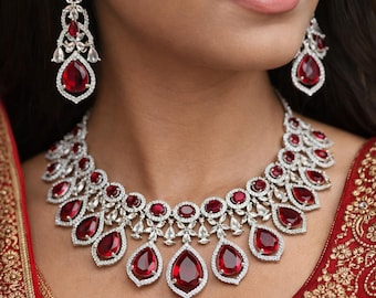 May include: A dazzling jewelry set featuring a necklace and matching earrings. The necklace and earrings showcase large, teardrop-shaped red gemstones, each surrounded by a halo of smaller, clear stones. The jewelry is set against a red fabric background.