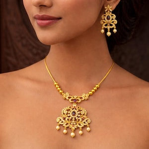 Op de afbeelding: Een gouden ketting en bijpassende oorbellen set. De ketting heeft een hanger met een centrale paarse steen, omringd door kleinere witte stenen en bungelende parels. De oorbellen hebben een vergelijkbaar ontwerp.