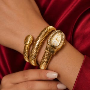 Op de afbeelding: Een goudkleurig horloge met een slangenarmband. De ovale wijzerplaat heeft Romeinse cijfers en een met diamanten bezette bezel. De armband wikkelt zich om de pols en creëert een uniek en stijlvol accessoire.