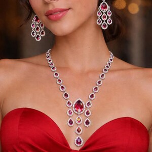 Peut inclure: Un ensemble de bijoux éblouissant comprenant un collier et des boucles d'oreilles assorties. Le collier présente un pendentif central avec une grosse pierre en forme de goutte de couleur rubis, entourée de plus petits bijoux. Les boucles d'oreilles reprennent le design du collier.