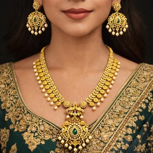 May include: A gold-toned necklace and matching earrings set. The necklace features a central pendant with a large green stone and pearl accents. The earrings have a similar design, with a green stone and dangling pearls. The jewelry is worn over a green and gold embroidered garment.