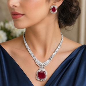 May include: A jewelry set consisting of a necklace and matching earrings. The necklace features a large, rectangular red gemstone pendant surrounded by small white stones, connected to a chain of white stones. The earrings match the pendant.