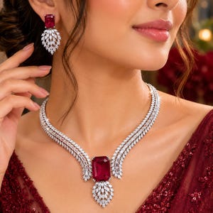 May include: A jewelry set consisting of a necklace and earrings. The necklace features a large rectangular red center stone flanked by rows of clear white stones, with a dangling leaf-shaped pendant. The earrings match with a red stone and leaf drop.