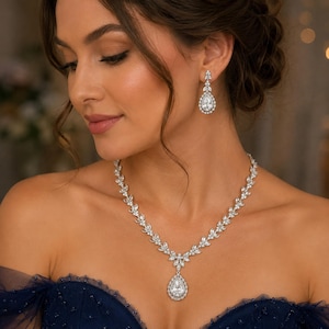 May include: A silver-toned necklace and matching drop earring set featuring clear, pear-shaped stones surrounded by smaller round stones. The necklace has a leaf-like vine pattern leading to a central pear-shaped pendant, and the earrings mirror the design.