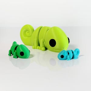 May include: Three 3D printed chameleon figurines. The largest chameleon is green and has a curled tail. The other two chameleons are smaller, one is green and the other is blue.