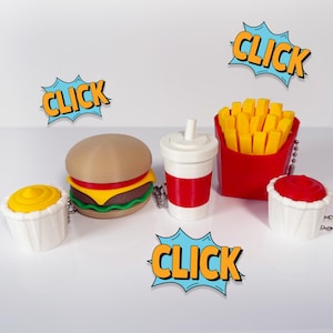 May include: A collection of fast food-themed keychains. Includes a hamburger, fries in a red carton, a soda, and small containers of yellow mustard and red ketchup. Each item has a silver chain. The word "CLICK" is in a comic book style.